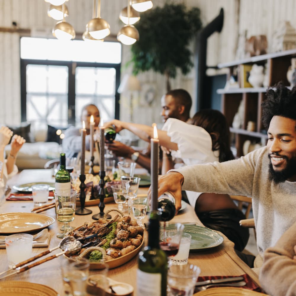 A group of friends enjoys a meal together around a warmly lit dining table, with two people pouring drinks, and various dishes and lit candles in a cozy, decorated room.