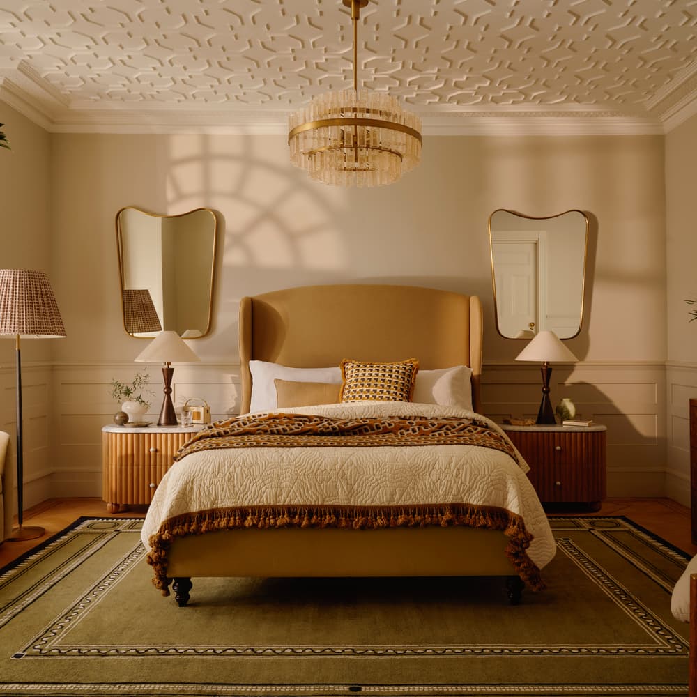 A neatly made bed stands in the center of a cozy bedroom, framed by two side tables, lamps, and large mirrors, with a chandelier above and decorative elements around.
