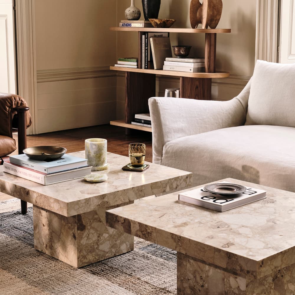 A modern living room with a white sofa and a brown leather chair surrounds two marble coffee tables holding books, candles, and a glass. A wooden shelf with various decor items is in the background.