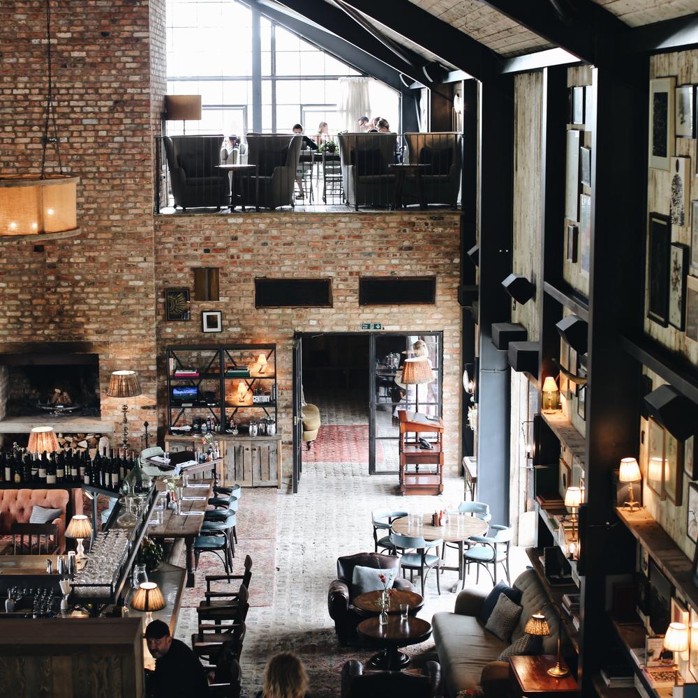 Brick-walled, loft-style bar and restaurant featuring industrial lighting, a cozy fireplace, and a central bar counter with seating. Patrons dine both upstairs and downstairs amidst shelves and framed wall art.
