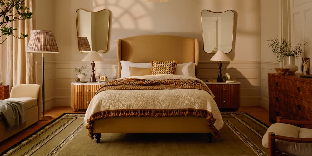 A bed with a beige headboard and decorative pillows is situated in a stylish bedroom, featuring matching nightstands, mirrored walls, a chandelier, an armchair, and a textured ceiling.