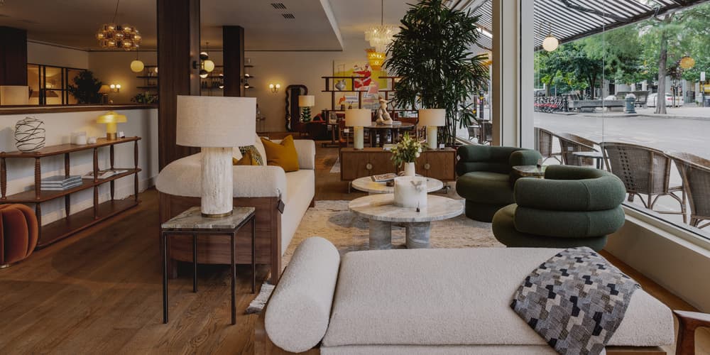 A cozy, modern showroom displays furniture, including sofas, lamps, and tables, with large glass windows revealing an outdoor patio and street view, adorned with plants and stylish decor elements.