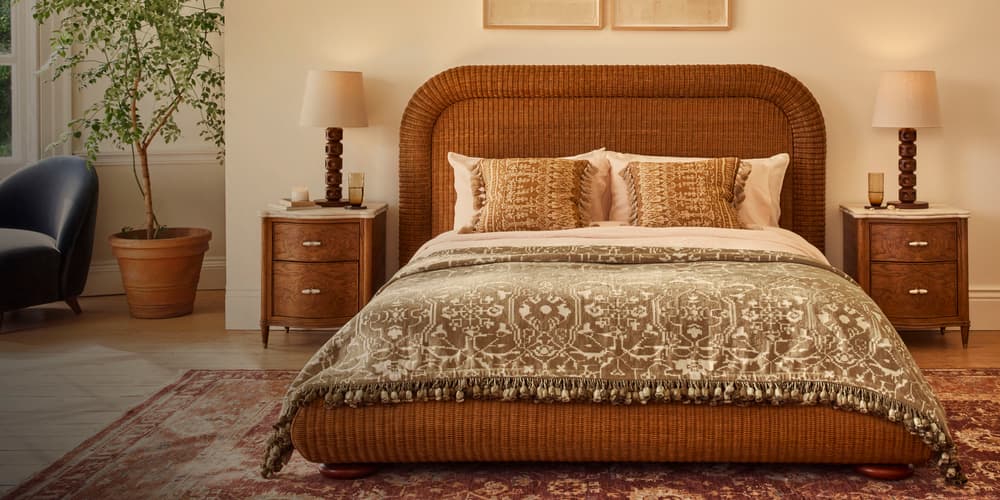 A wicker bed, adorned with patterned pillows and a blanket, is positioned centrally. Flanked by wooden nightstands and lamps, a large potted plant and an armchair decorate the room.