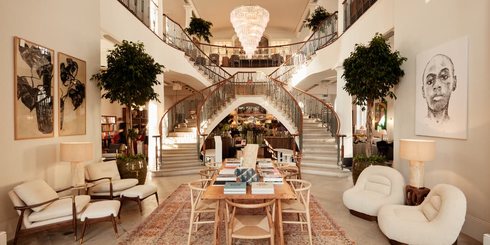 A wooden table with books and chairs sits on a rug in a luxurious room featuring a grand double staircase, large artwork, lush plants, and elegant seating.