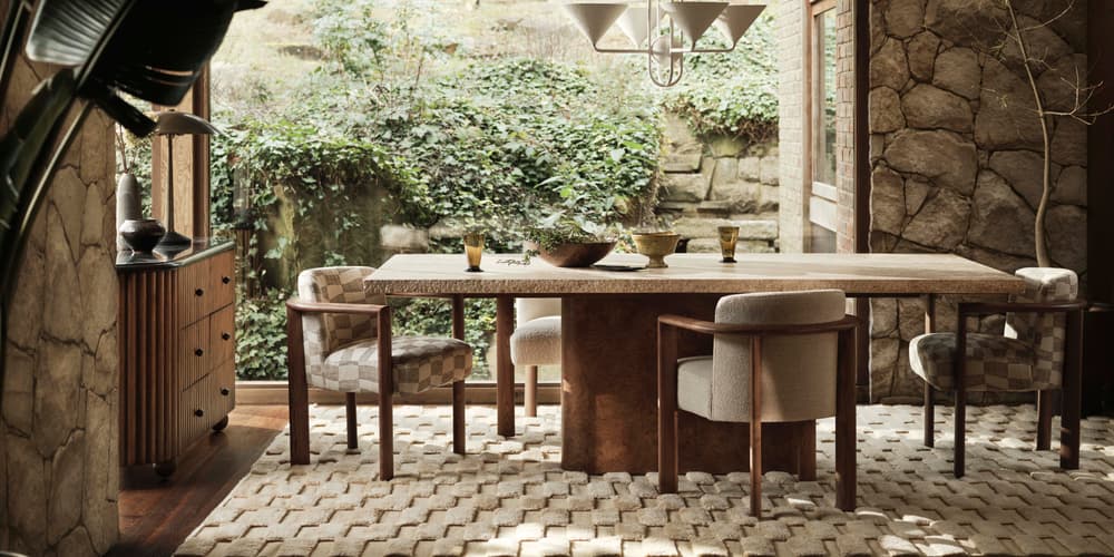 A dining table surrounded by cushioned chairs with a pendant light overhead in a stone-walled room, featuring large windows, green foliage outside, a patterned rug, and wooden furniture.