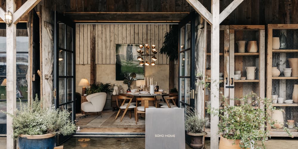 A cozy showroom features a dining table setting with chairs, surrounded by rustic decor. Potted plants and pottery sit outside; a sign reads "SOHO HOME."
