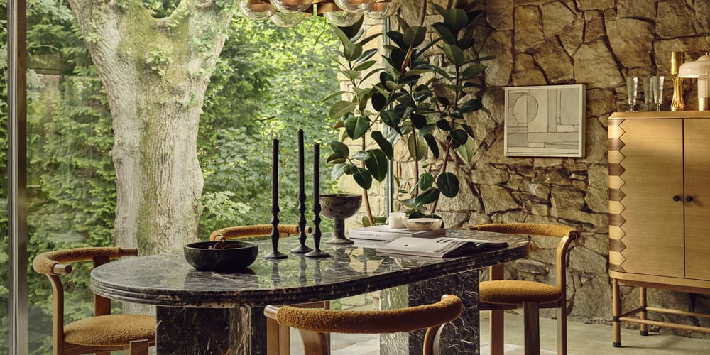 A marble dining table with four chairs stands in a room with stone walls, large windows showing a tree, and a wooden cabinet; black candlesticks and a bowl sit on the table.