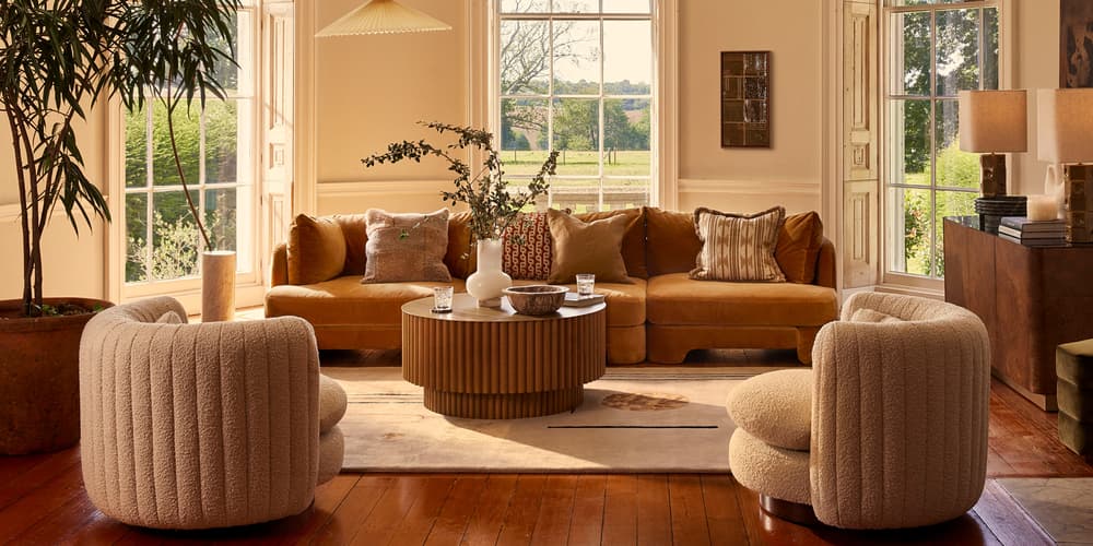 A mustard-colored sectional sofa and two beige armchairs surround a wooden coffee table with vases and glasses in a bright living room, featuring tall windows, a chandelier, and plants.