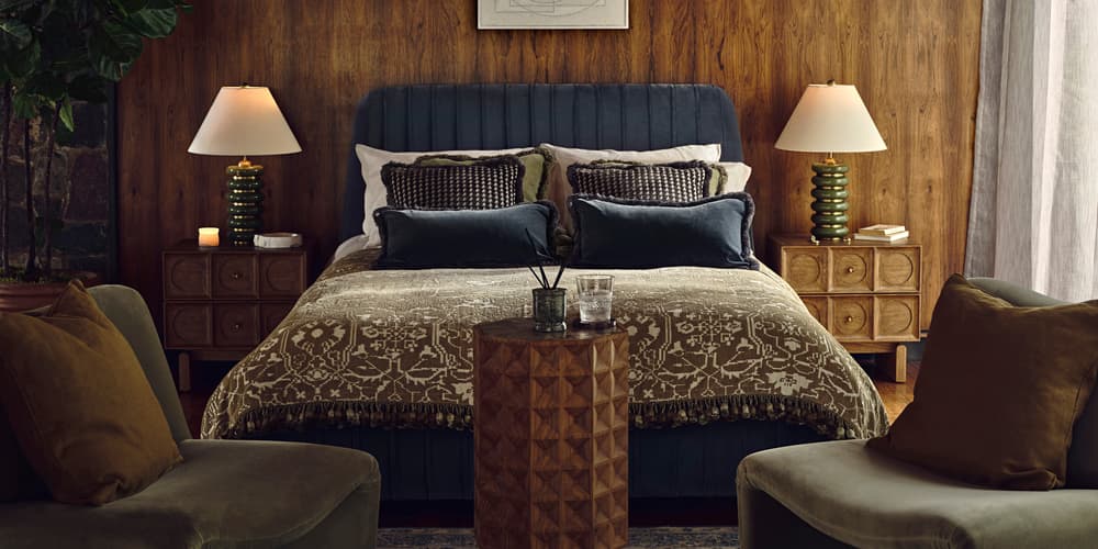 A double bed with patterned bedding and pillows sits against a wood-paneled wall, flanked by two lamps on bedside tables. Two chairs and a small table are in the foreground.