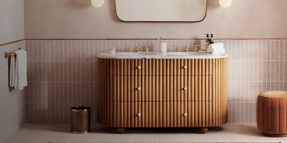 Wooden bathroom vanity with a marble countertop, under a wavy-edged mirror, flanked by round wall lights and a hand towel on a rack, set against tiled and textured walls.