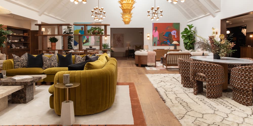 A large living room features a mustard-yellow velvet couch with patterned cushions, surrounded by modern decor, artwork, plants, wooden furniture, and hanging light fixtures under a vaulted, white ceiling.