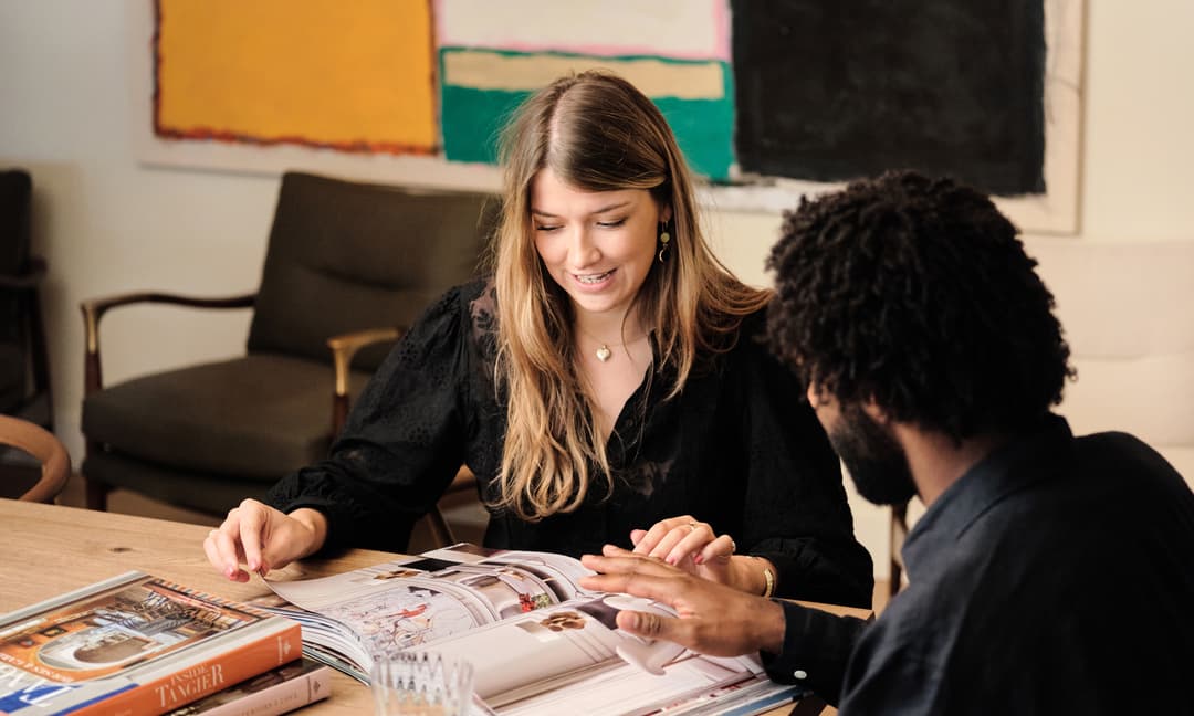 Two individuals are seated at a wooden table, reviewing a magazine together. They are in a room with chairs and a large colorful abstract painting on the wall behind them.