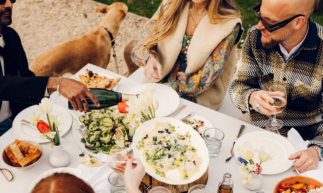 People are enjoying an outdoor meal at a white table, adorned with salads, bread, and drinks. A man pours champagne, and a dog is in the background near a garden.
