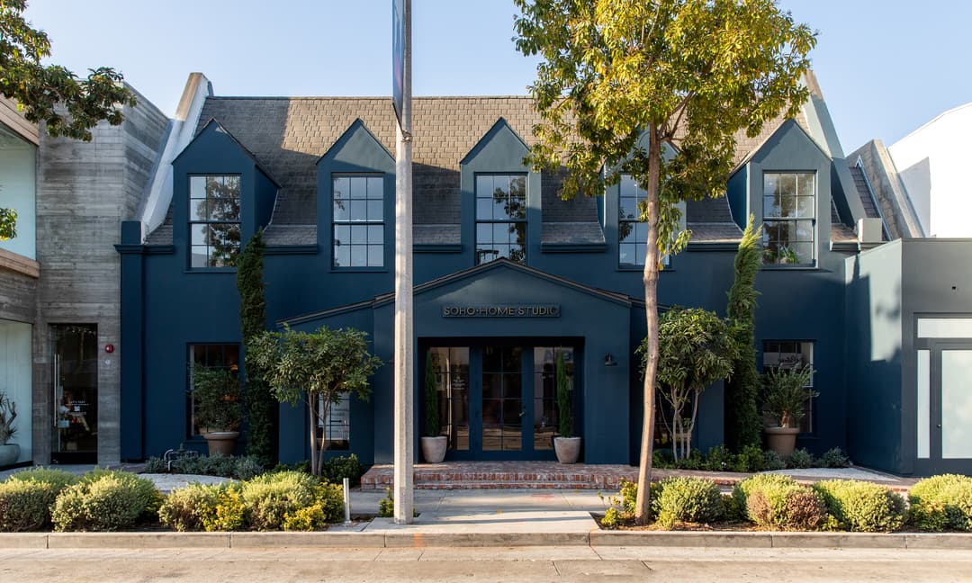 A blue-gray two-story building with large windows and a sign reading "SOHO HOME STUDIO" above the entrance, surrounded by trees and shrubs on both sides along a quiet street.