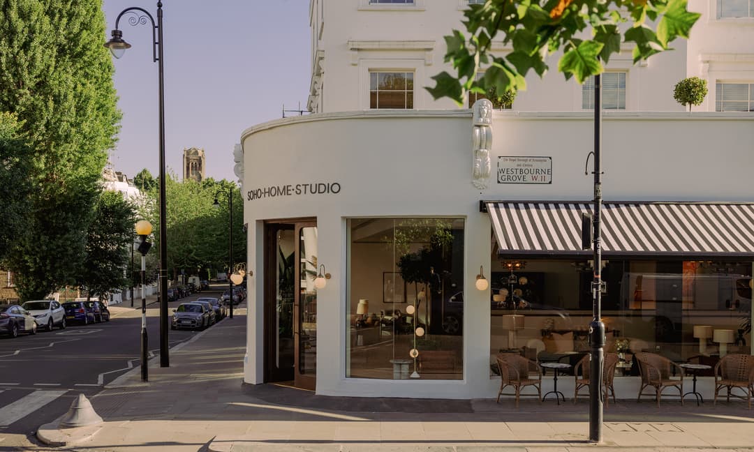 A corner building labeled "SOHO HOME STUDIO," with striped awning, large windows, outdoor seating, and street signs reading "Westbourne Grove W.11," on a sunny street lined with cars and trees.