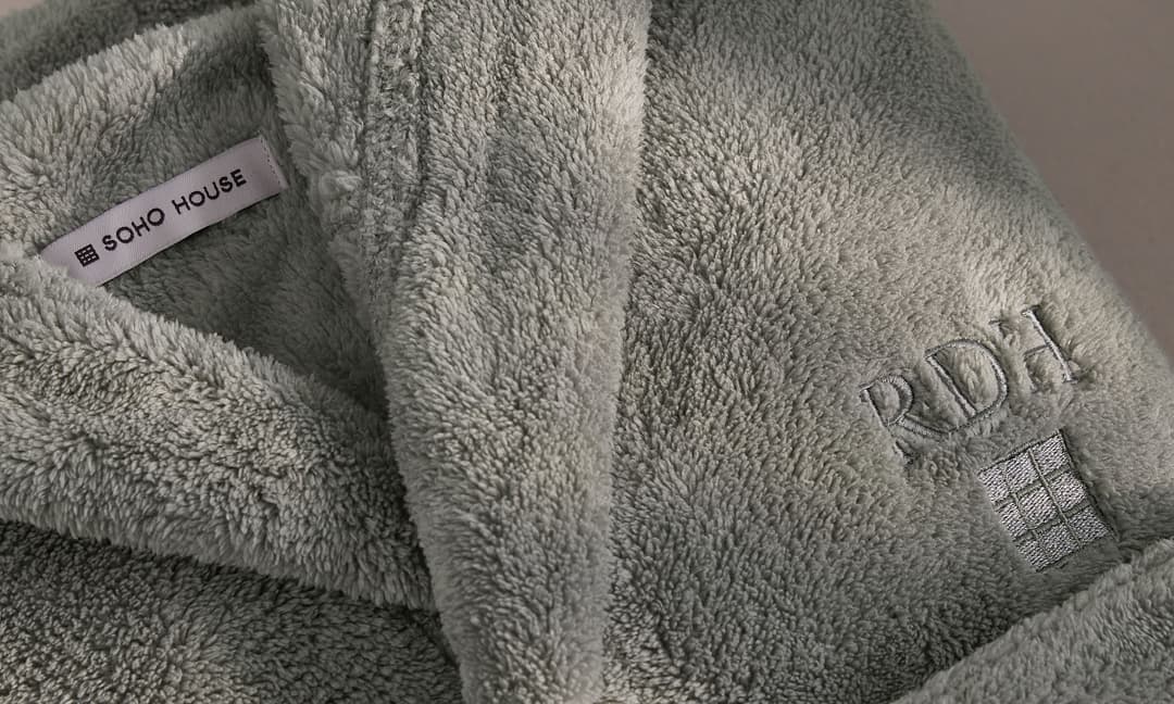 A folded, plush gray robe shows a label reading "SOHO HOUSE" inside the collar and an embroidered monogram "RDH" with a small window icon on the exterior.