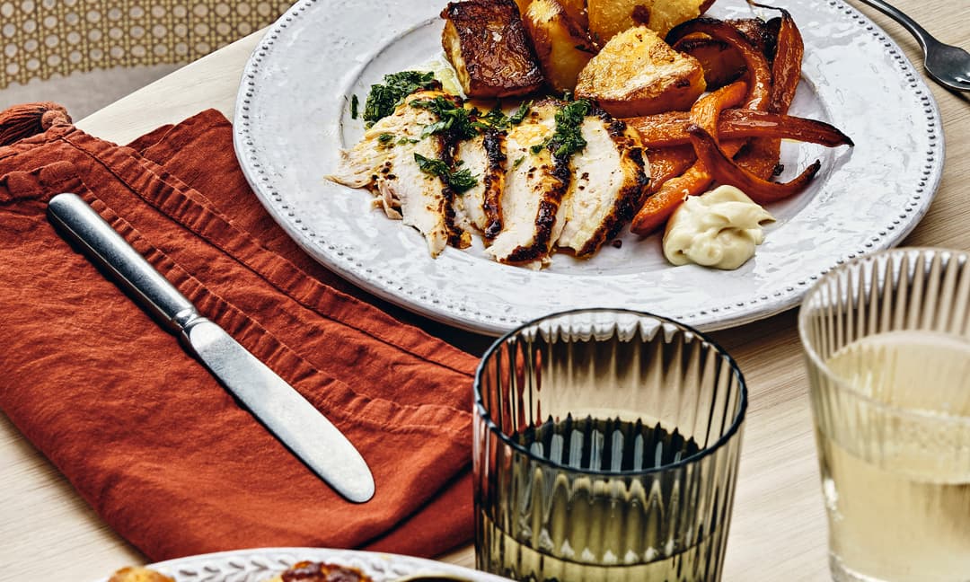Dinner plate with sliced chicken, roast potatoes, and carrots. Nearby, a glass of wine, a black-handled knife on a red napkin, and wooden chair at a wooden dining table.