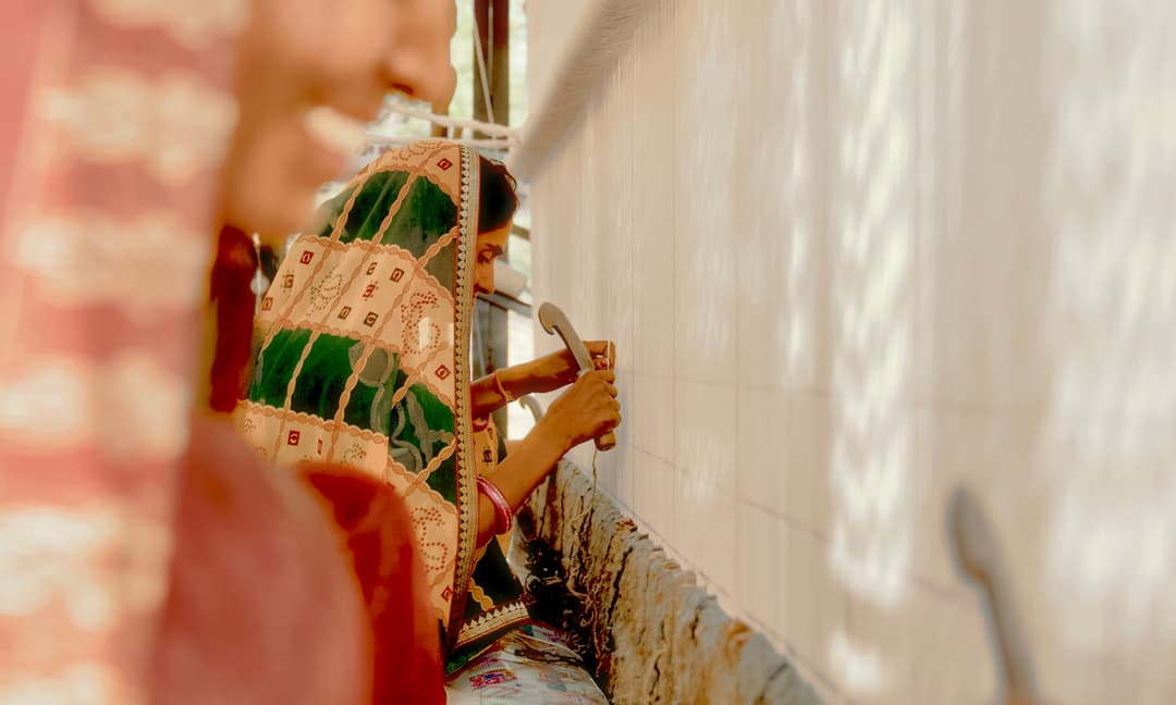 Two women, dressed in colorful, patterned fabrics, meticulously tie knots on a large, vertical tapestry loom in a sunlit room. One woman's face is blurred in the foreground.