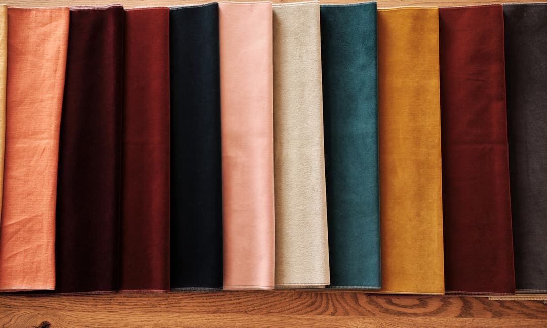 Folded fabric swatches are arranged in a row, showcasing different colors including peach, burgundy, beige, teal, mustard, and brown hues on a wooden surface.