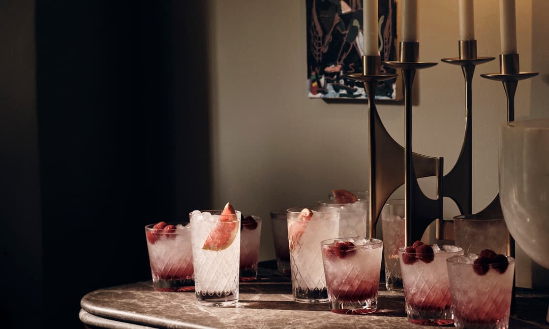 Eight iced cocktails containing berries or fruit garnish rest on a marble countertop beside a golden candlestick in a softly lit room with a framed artwork on the wall.