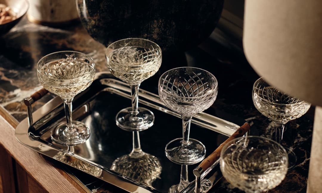 Five crystal glasses with intricate patterns rest on a polished silver tray. Each glass holds a light-colored liquid. The tray is placed on a dark marble surface with various decorative items around.