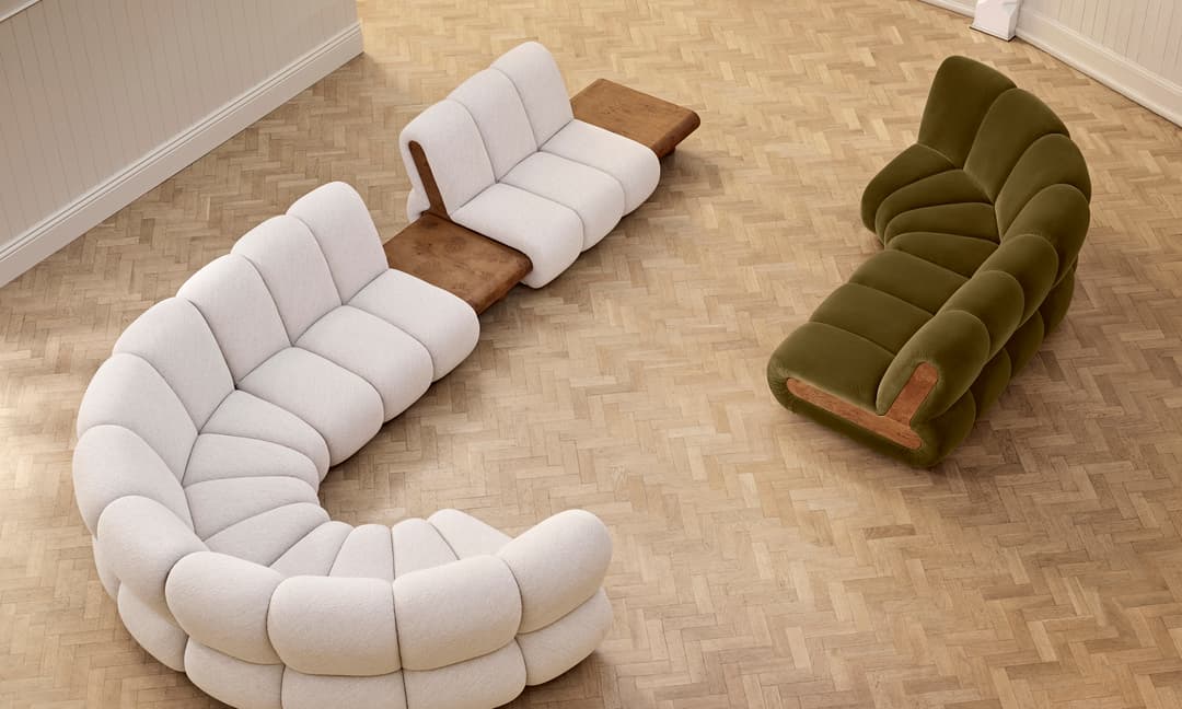 A cream modular sectional sofa with a wooden side table is positioned in a curved arrangement on a herringbone-patterned floor, facing an olive-green sofa in a minimalist room.