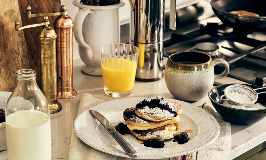 Pancakes topped with blueberries and powdered sugar, placed on a white plate with a knife, surrounded by a bottle of milk, a mug, orange juice, and cooking utensils on a kitchen counter.