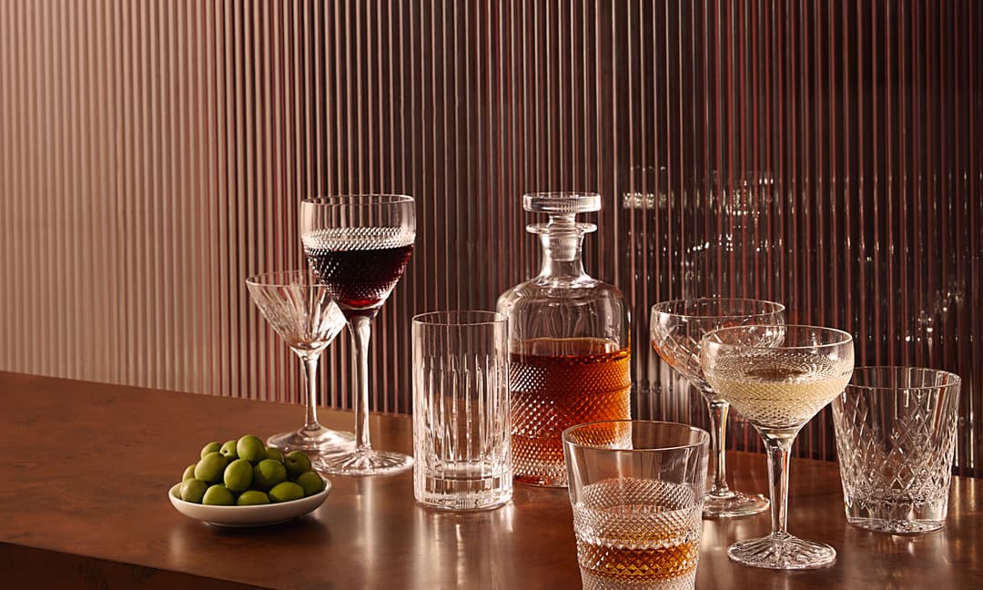 Various filled crystal glasses and a decanter are arranged on a wooden surface, accompanied by a small bowl of green olives, set against a vertically striped, translucent background.