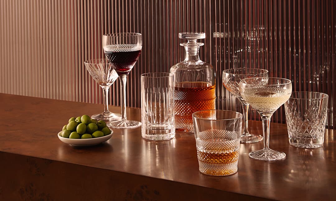 Various filled crystal glasses and a decanter are arranged on a wooden surface, accompanied by a small bowl of green olives, set against a vertically striped, translucent background.