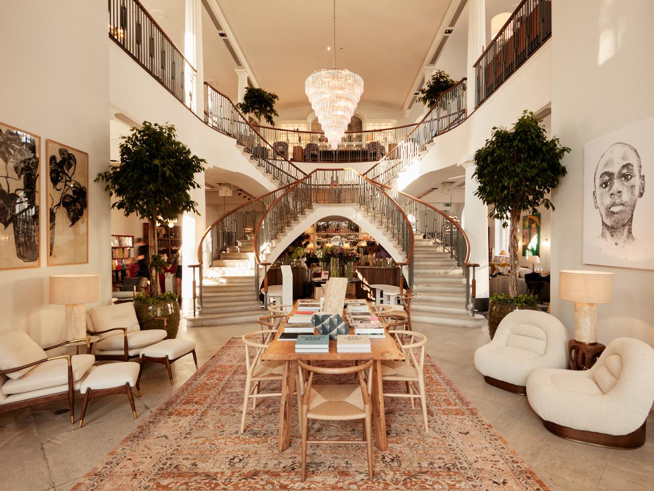 Grand staircase in a luxurious, modern space with elegant furnishings. Two large plants flank a central wooden table with chairs on an ornate rug. Artwork and seating adorn the walls.