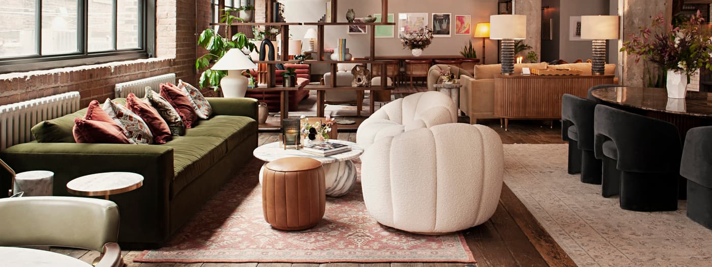 Modern lounge with a green sofa, patterned cushions, and a white armchair, featuring a marble coffee table and leather stool, all set in an industrial loft with large windows and hanging chandeliers.