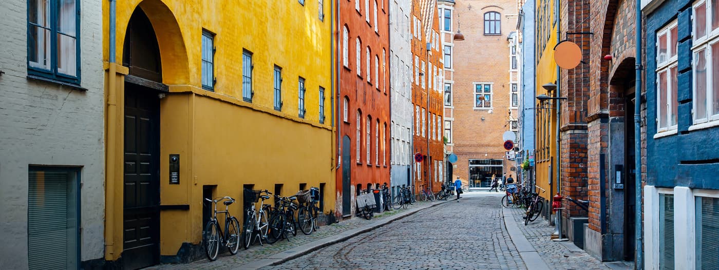 Bicycles are parked along a narrow, cobblestone street lined with colorful buildings in shades of yellow, orange, and brick red, in a quaint, European cityscape setting.