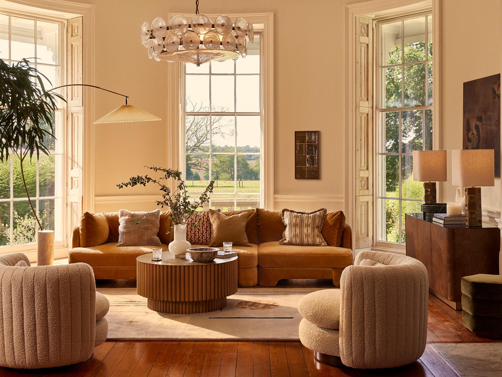 A mustard-colored sectional sofa and two beige armchairs surround a wooden coffee table with vases and glasses in a bright living room, featuring tall windows, a chandelier, and plants.