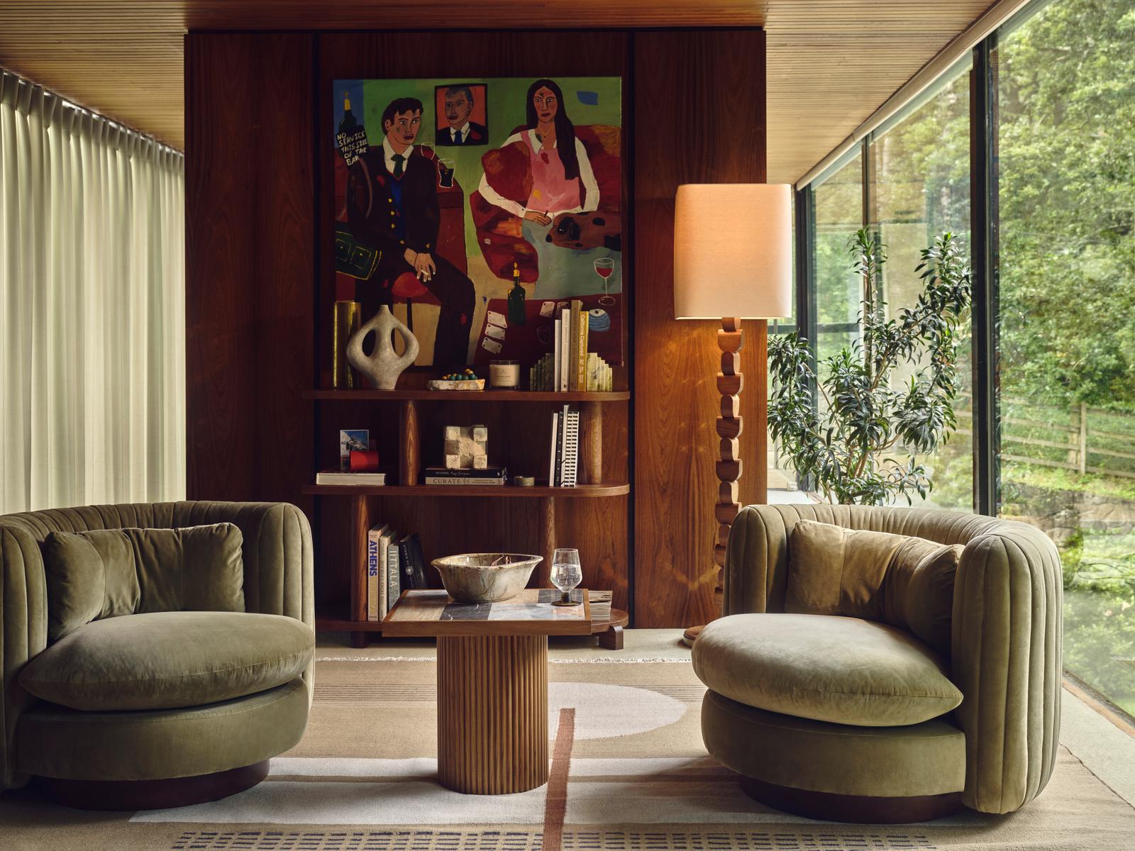 Two round velvet chairs face a wooden coffee table in a cozy living room with bookshelves, a modern artwork, large windows, and lush greenery outside. A unique floor lamp stands beside the chairs.