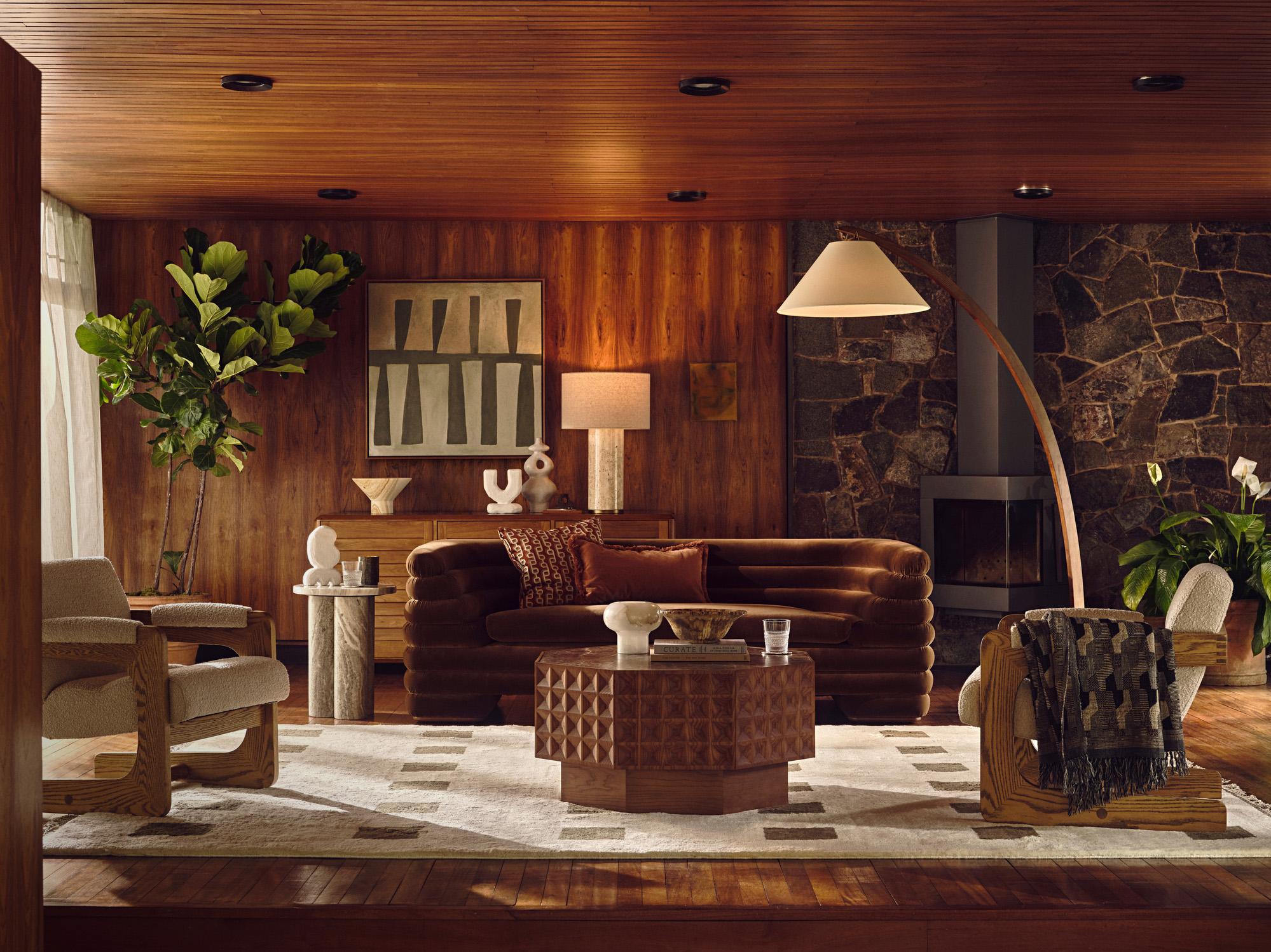 Living room featuring a brown velvet sofa, wooden armchairs, and a brown geometric coffee table holding décor items. Wood-paneled walls, stone fireplace, large floor plant, and abstract wall art enhance the cozy, elegant atmosphere.