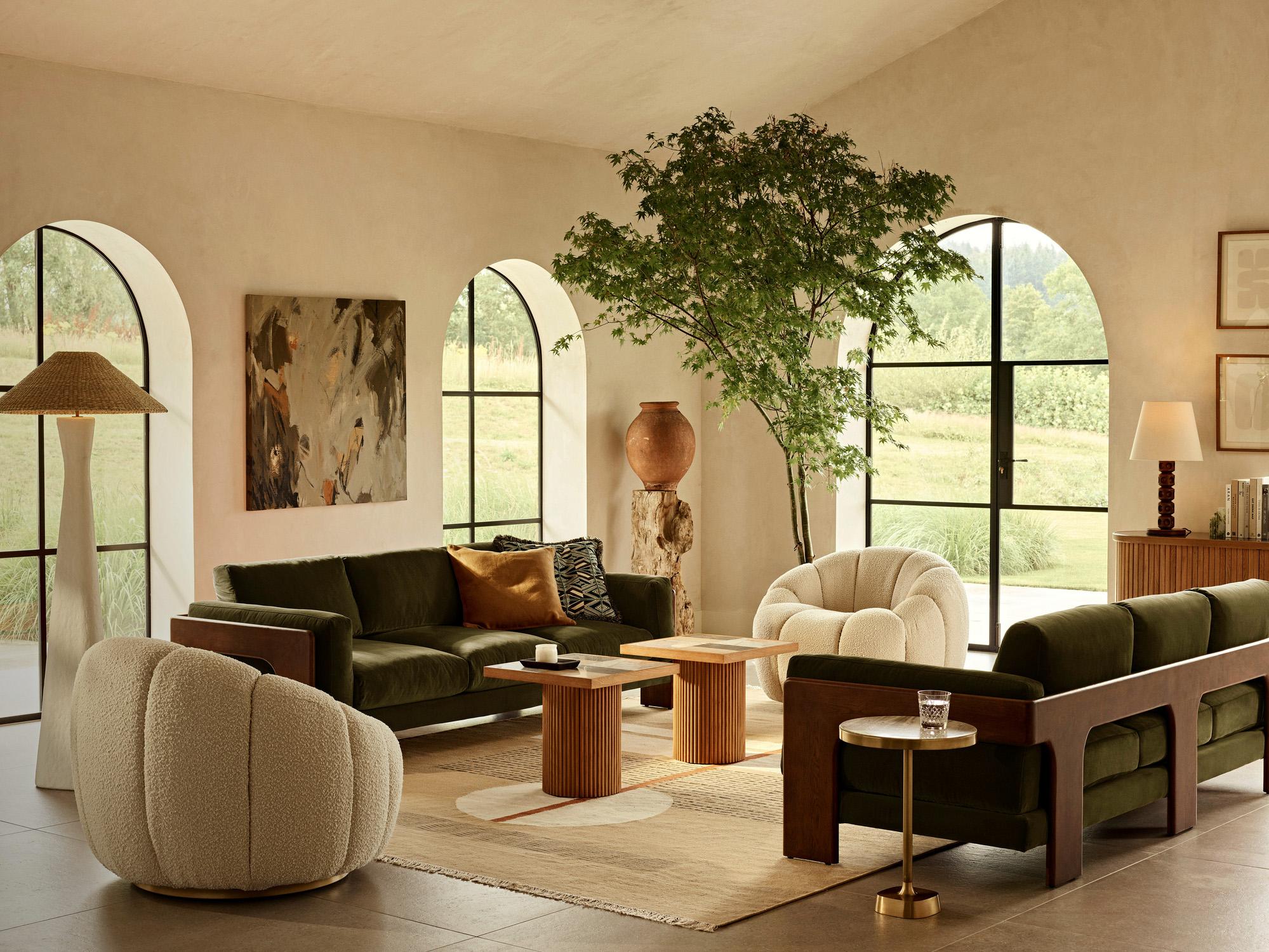 A cozy living room with plush green and beige chairs encircling low wooden tables. Large arched windows reveal a green landscape, and indoor plants and modern lamps enhance the serene atmosphere.