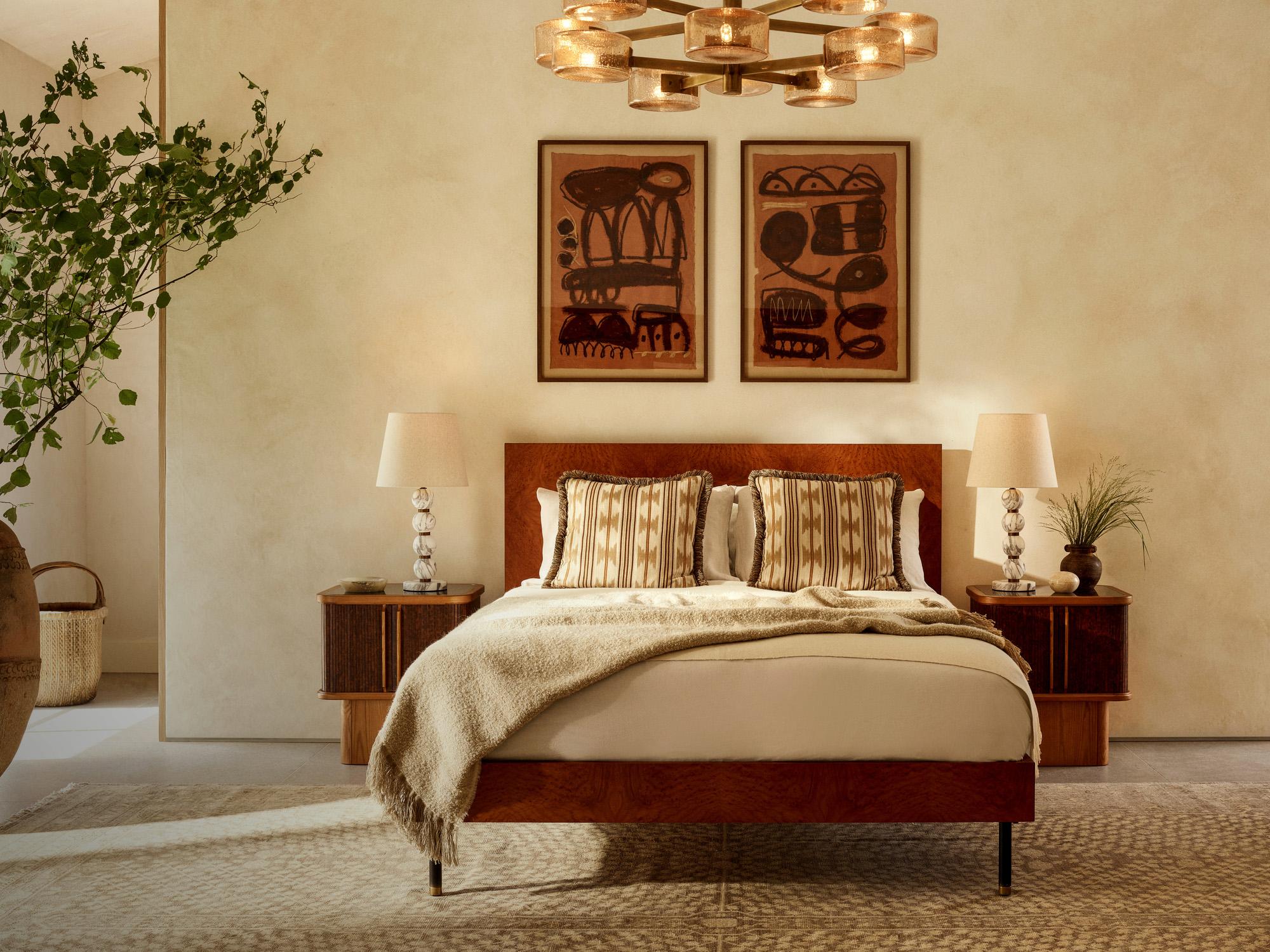 A bed with patterned pillows and a beige throw rests between two nightstands and lamps; framed abstract art and a large plant adorn the softly lit bedroom.