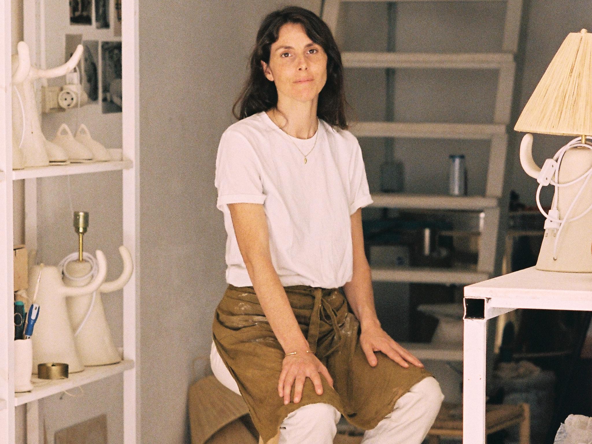 A woman sits on a wooden stool, wearing a white shirt and brown apron, in a cluttered studio with shelves holding various ceramics and art supplies. The background reveals a loft space with a ladder.