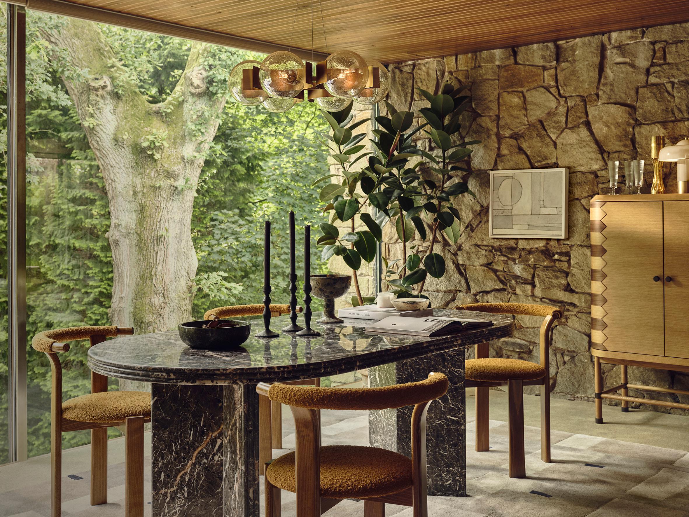 A marble dining table with four chairs stands in a room with stone walls, large windows showing a tree, and a wooden cabinet; black candlesticks and a bowl sit on the table.