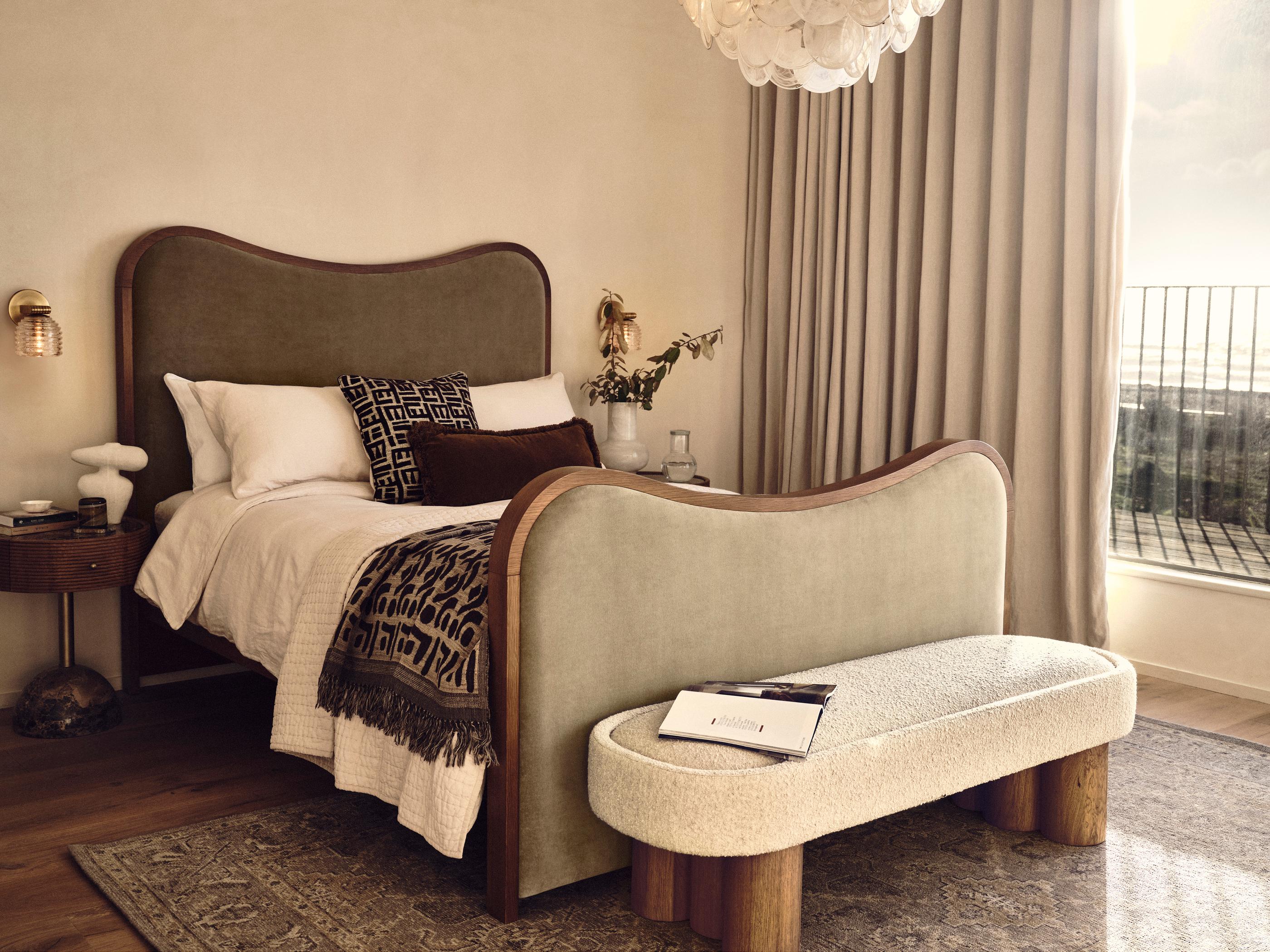 A neatly made bed with plush pillows and a blanket sits in a cozy bedroom adorned with a side table, bench, chandelier, and large window with curtains overlooking a balcony.