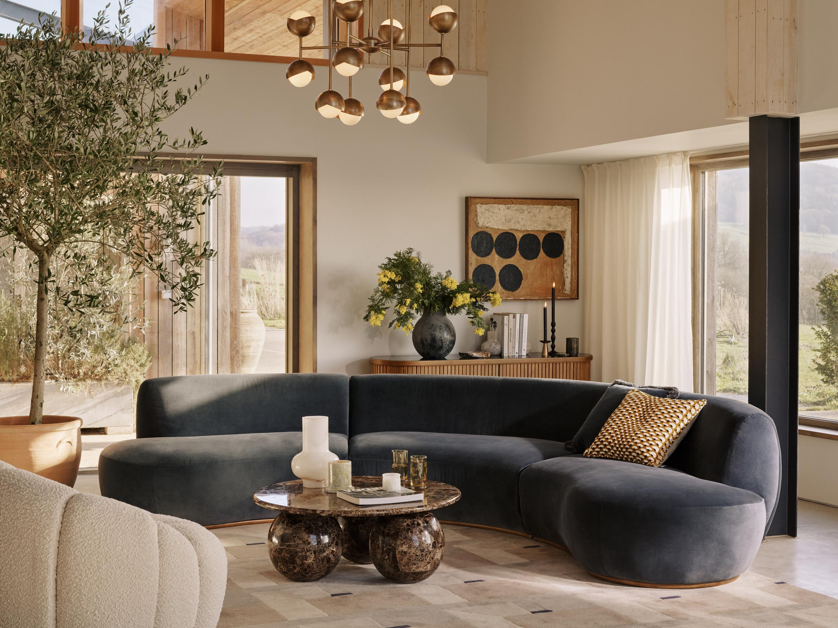 A curved dark gray sofa sits in a sunlit living room with beige walls, large windows, and a beige armchair; next to a marble coffee table holding decorative items. A potted tree, framed art, and flowers embellish the space.