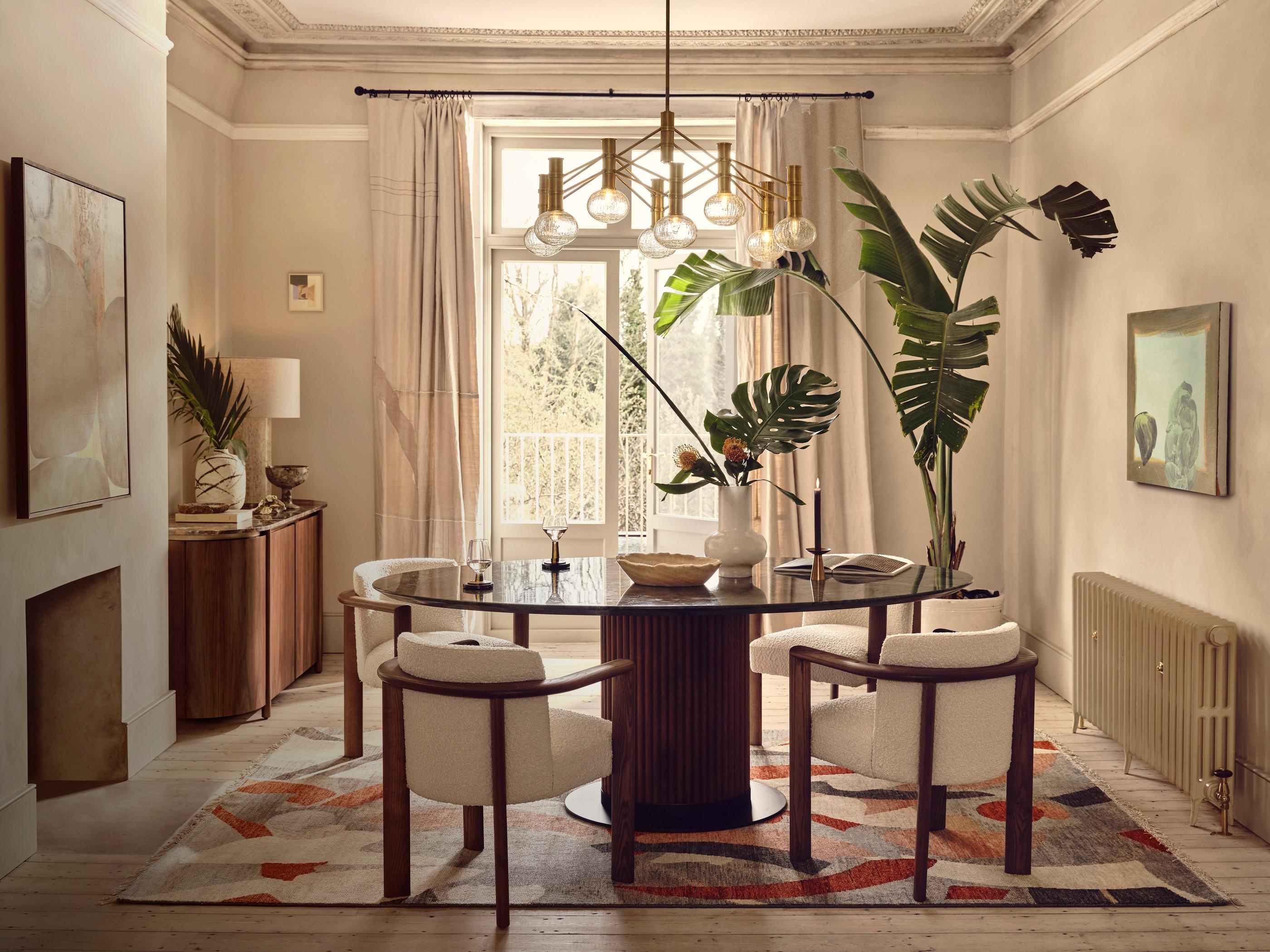 A round wooden dining table with chairs, adorned with a centerpiece, set in an elegant room with large windows, indoor plants, wall art, a sideboard, and a patterned rug.