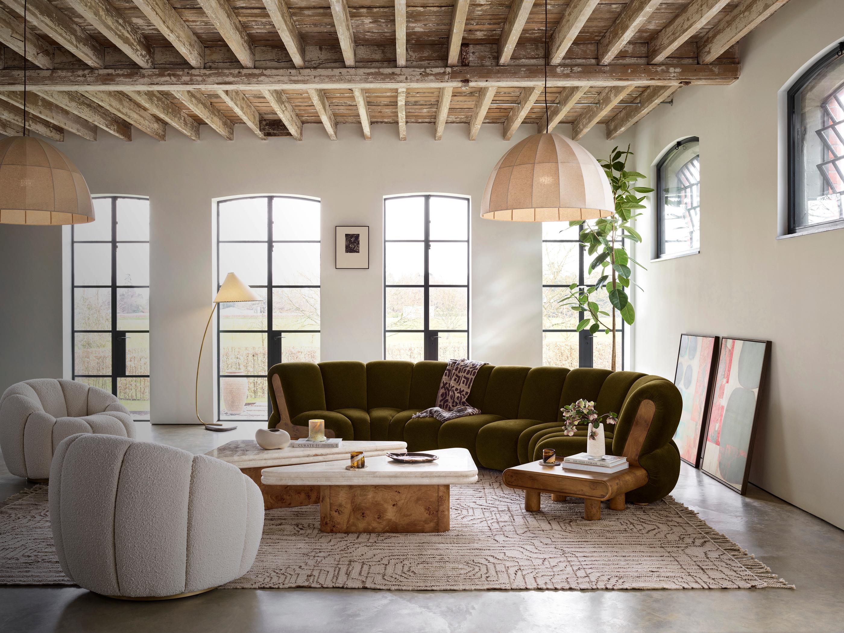 A green sectional sofa and two white plush chairs are arranged around wooden coffee tables in a spacious living room featuring large windows, a rustic wooden ceiling, and modern decor items.