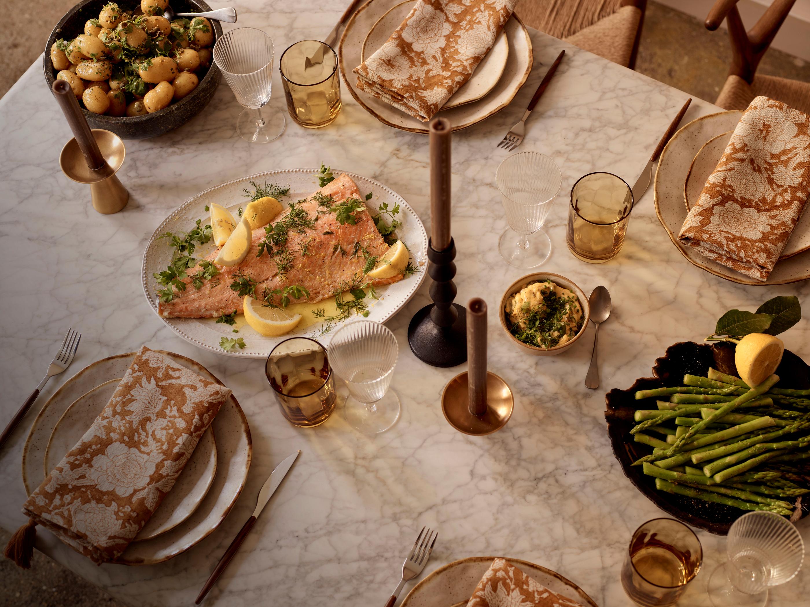 A marble table set for dining with plates, gold-patterned napkins, glasses, and food: a platter with cooked salmon and lemon, a bowl of asparagus, and another with potatoes.