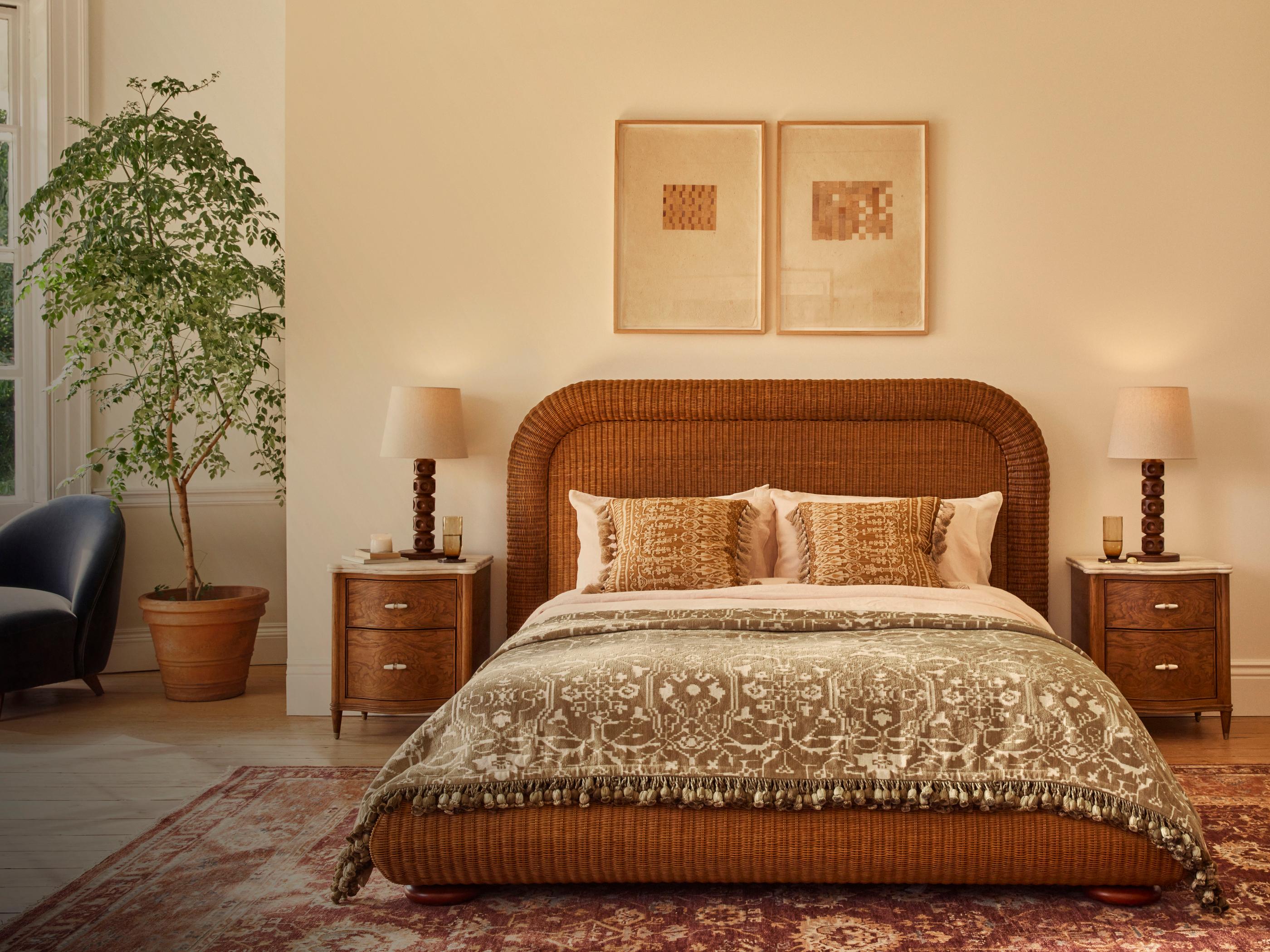 A wicker bed, adorned with patterned pillows and a blanket, is positioned centrally. Flanked by wooden nightstands and lamps, a large potted plant and an armchair decorate the room.