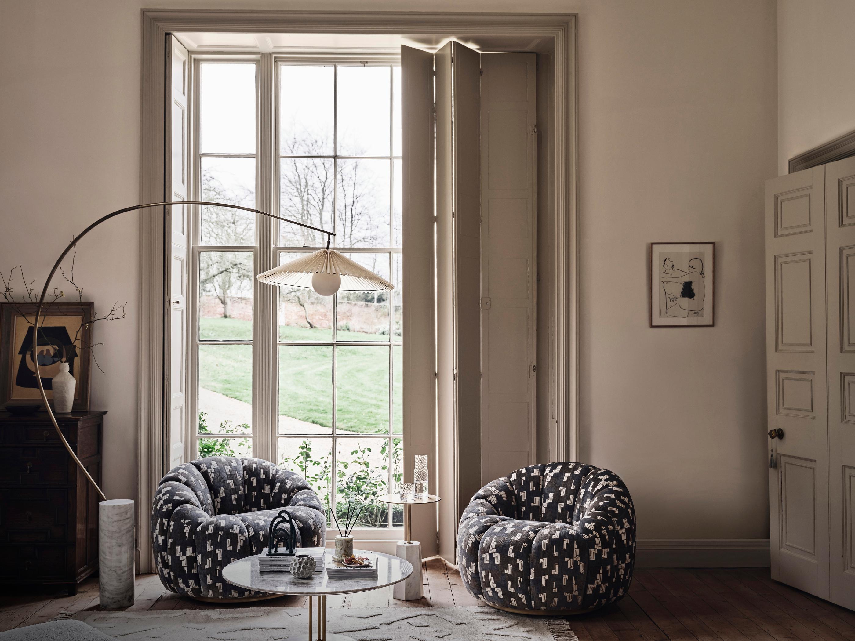 Two cushy, patterned chairs, flanking a small side table with decorative items, are positioned in front of large, open, paneled windows in a softly lit, elegant living room.