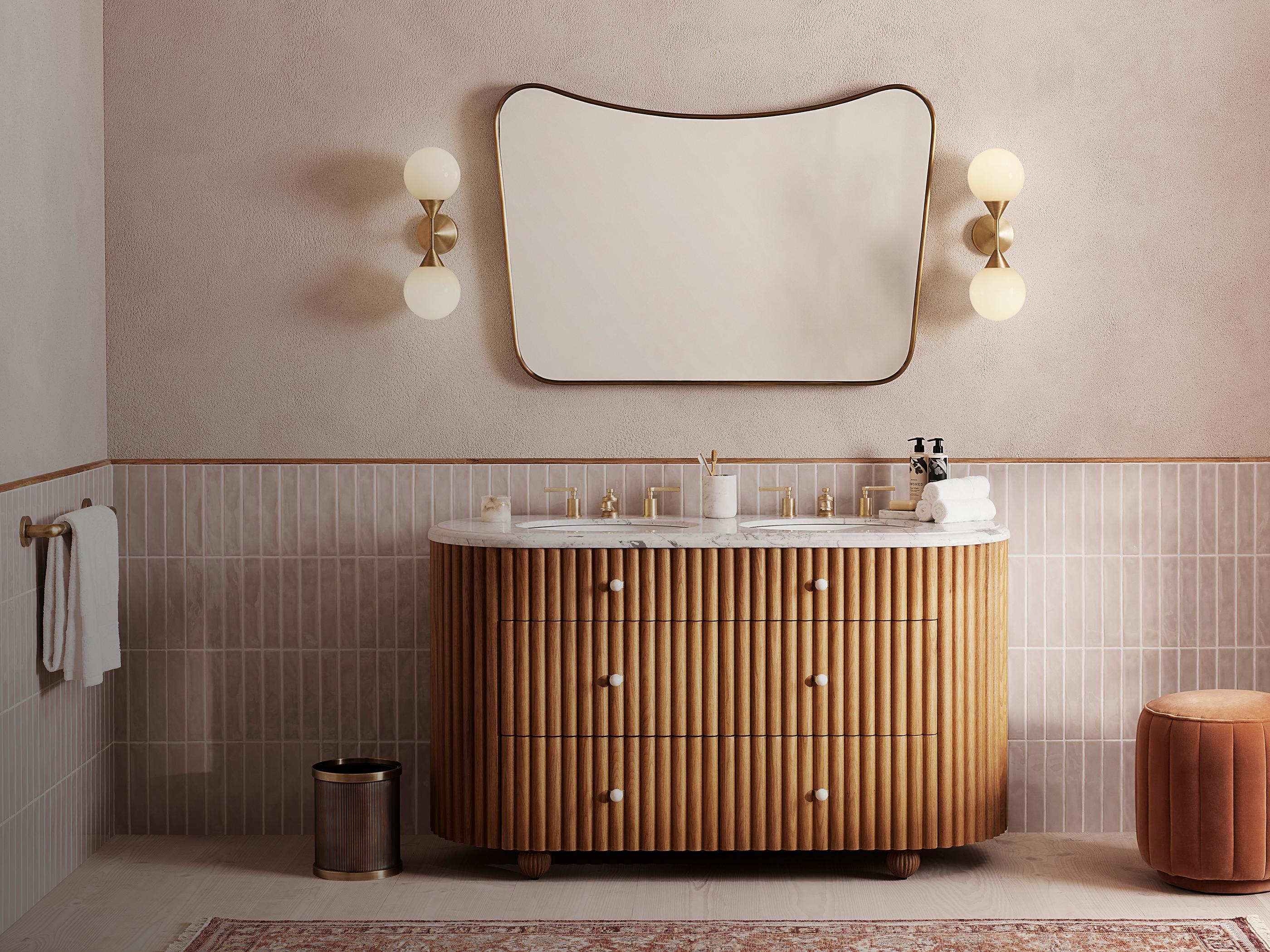 Wooden bathroom vanity with a marble countertop, under a wavy-edged mirror, flanked by round wall lights and a hand towel on a rack, set against tiled and textured walls.