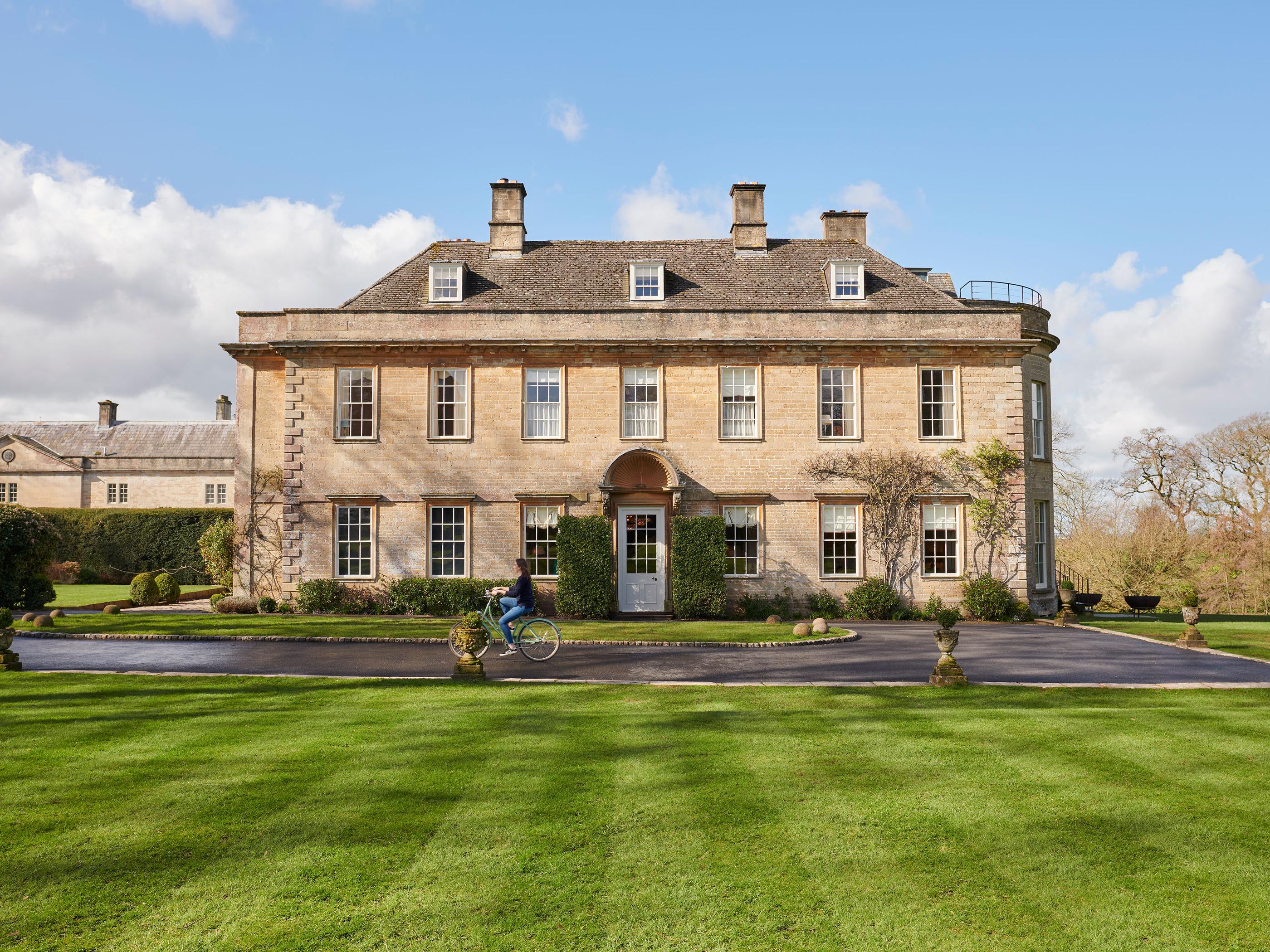 A person rides a bicycle past a large, symmetrical, three-story stone mansion with multiple windows in a landscaped garden with neatly trimmed grass and hedges under a bright blue sky.