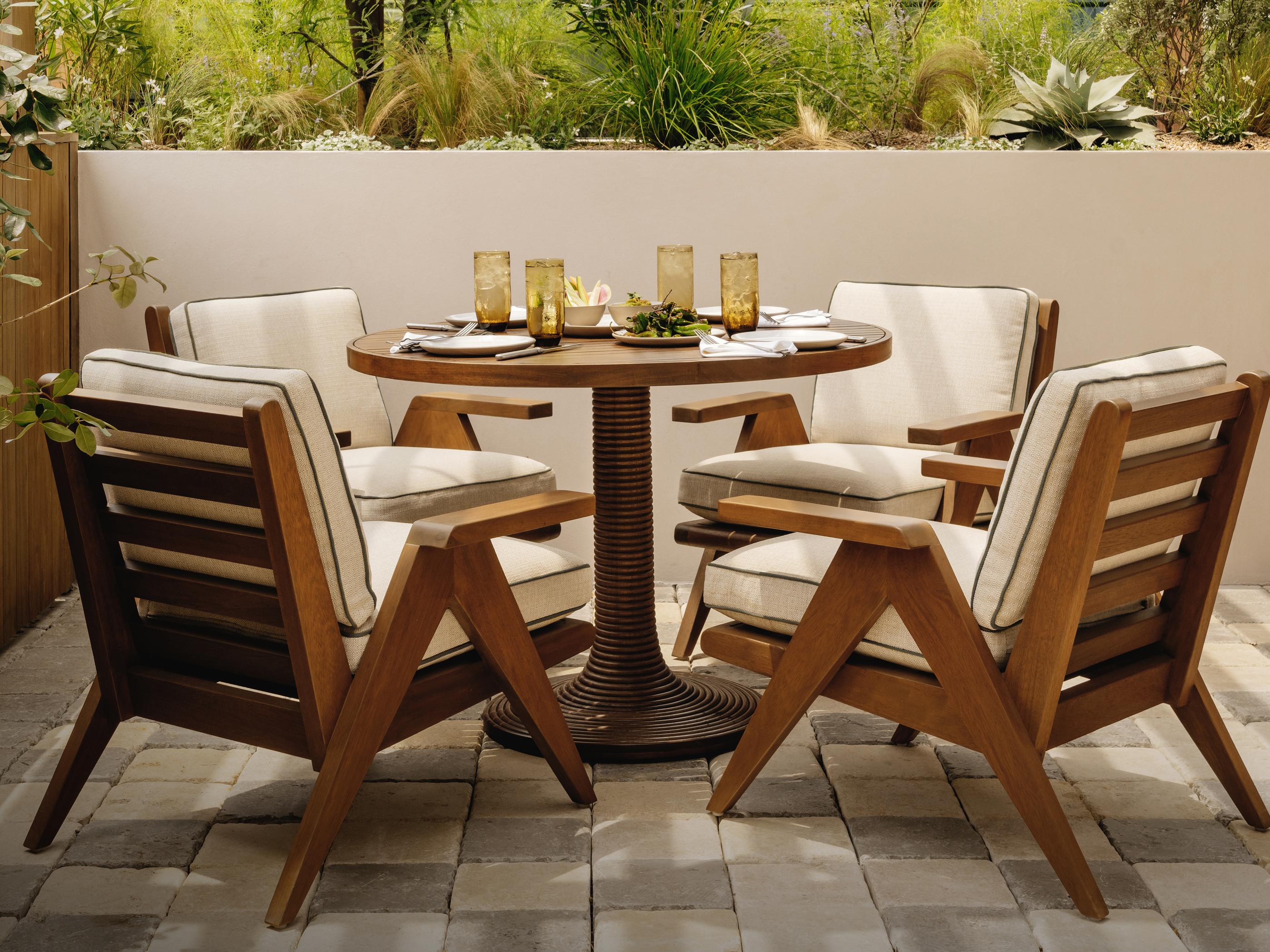 Four wooden chairs with beige cushions surround a round wooden table set with napkins, utensils, and amber-colored glasses on a stone patio with lush greenery in the background.