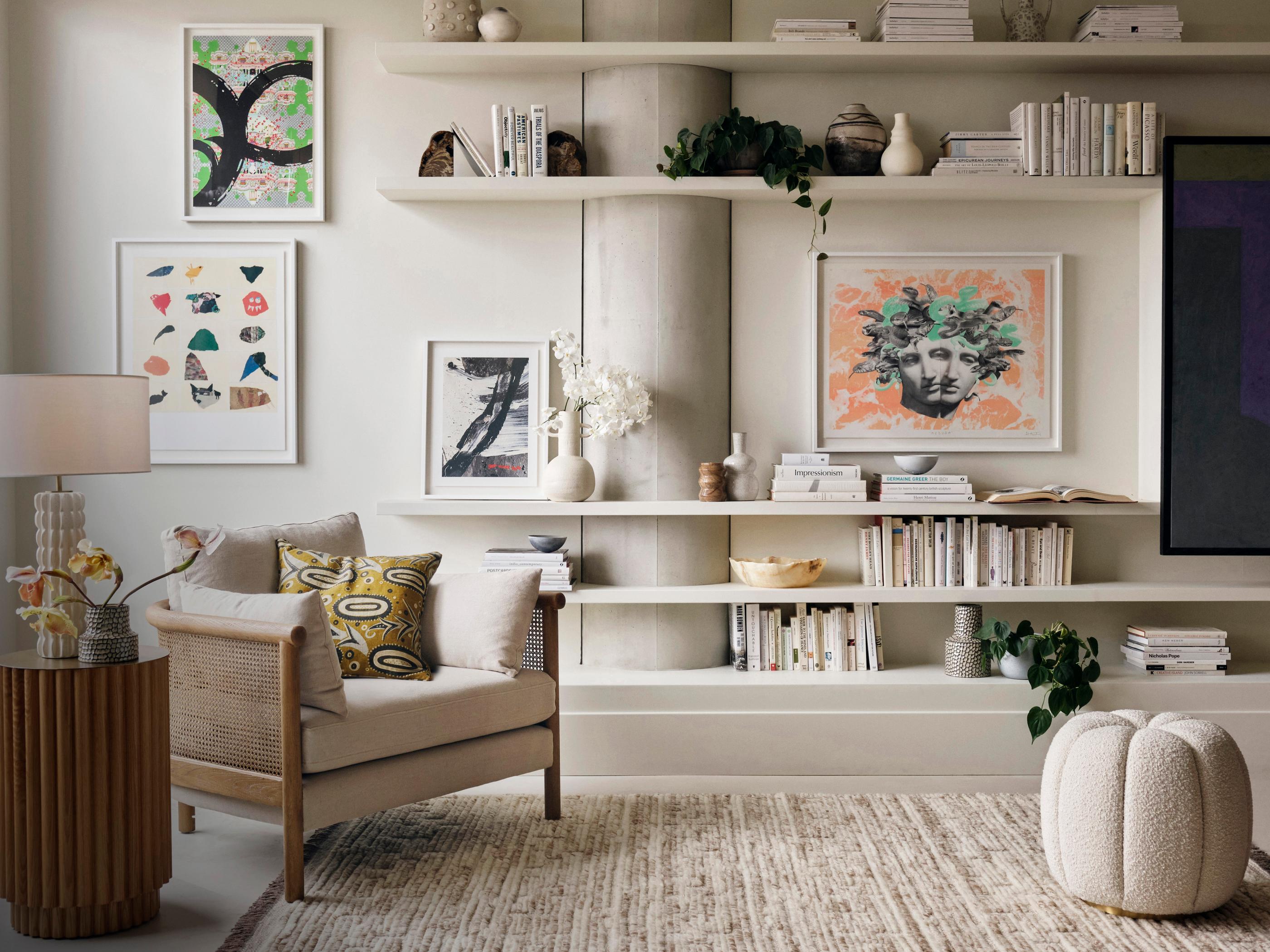 An armchair with cushions sits beside a small table with a lamp and vase; framed artwork and books on shelves decorate the cozy, modern living room with neutral colors.